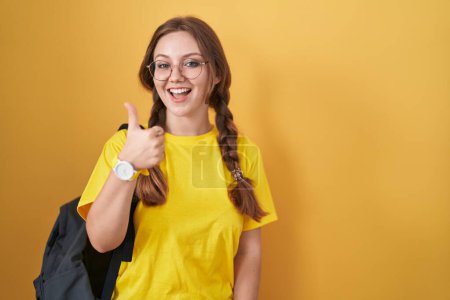 Téléchargez les photos : Jeune femme caucasienne portant un sac à dos étudiant sur fond jaune faisant des pouces heureux geste avec la main. approuvant l'expression regardant la caméra montrant le succès. - en image libre de droit