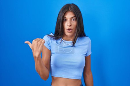 Photo for Brunette young woman standing over blue background surprised pointing with hand finger to the side, open mouth amazed expression. - Royalty Free Image