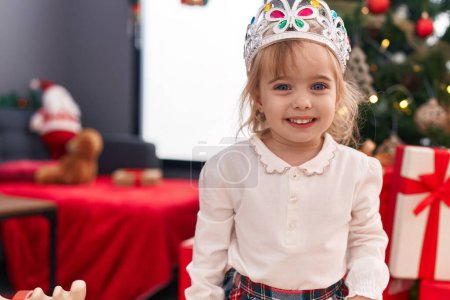 Foto de Adorable chica rubia con corona de princesa de pie junto al árbol de Navidad en casa - Imagen libre de derechos