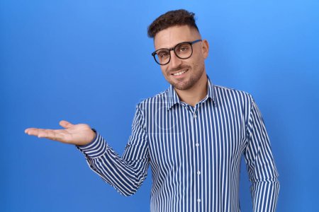 Photo for Hispanic man with beard wearing glasses smiling cheerful presenting and pointing with palm of hand looking at the camera. - Royalty Free Image