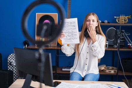 Téléchargez les photos : Jeune femme caucasienne faisant tutoriel de musique en ligne montrant feuille de musique couvrant la bouche avec la main, choquée et effrayée par l'erreur. expression surprise - en image libre de droit