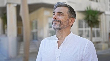 Foto de Joven hombre hispano confiado, de pelo gris, radiante y mirando al cielo, parado en la soleada calle de la ciudad con telón de fondo urbano, emitiendo alegría y un ambiente positivo. - Imagen libre de derechos