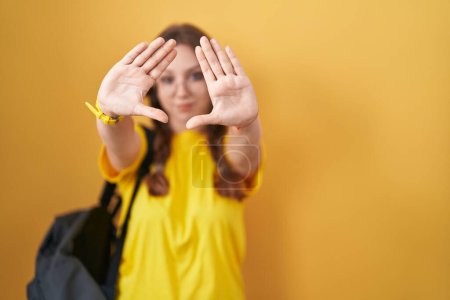Téléchargez les photos : Jeune femme caucasienne portant sac à dos étudiant sur fond jaune faisant cadre à l'aide de mains paumes et doigts, perspective de la caméra - en image libre de droit
