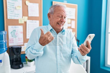 Téléchargez les photos : Homme âgé avec les cheveux gris travaillant au bureau faire appel vidéo avec smartphone pointant le pouce vers le côté souriant heureux avec la bouche ouverte - en image libre de droit