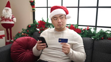 Photo for Young chinese man using smartphone and credit card sitting on sofa by christmas decoration at home - Royalty Free Image