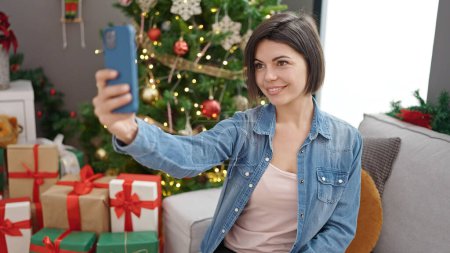 Téléchargez les photos : Jeune femme caucasienne prenant selfie avec smartphone debout près de l'arbre de Noël à la maison - en image libre de droit