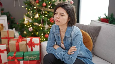 Photo for Young caucasian woman sitting on sofa by christmas tree looking upset at home - Royalty Free Image