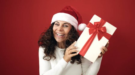 Téléchargez les photos : Moyen Age femme hispanique souriant confiant tenant cadeau de Noël sur fond rouge isolé - en image libre de droit