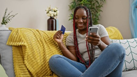 Foto de Confiada hermosa mujer afroamericana disfruta de compras en línea desde su sala de estar, sonriendo mientras usa su teléfono inteligente y tarjeta de crédito mientras está sentada en el sofá en casa - Imagen libre de derechos