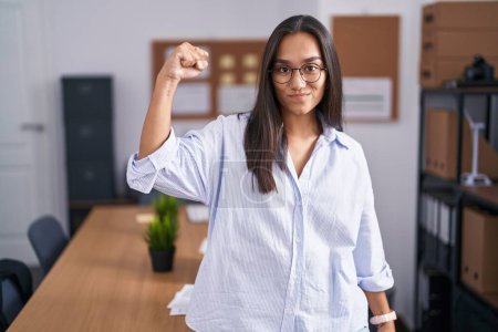 Téléchargez les photos : Jeune femme hispanique au bureau personne forte montrant muscle du bras, confiant et fier de la puissance - en image libre de droit