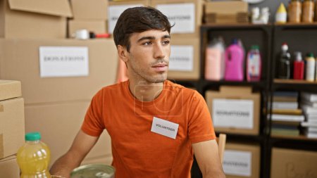 Photo for Devoted young hispanic man actively volunteering at a charity center, sitting at the table with a serious face, working diligently in a room full of donations - Royalty Free Image