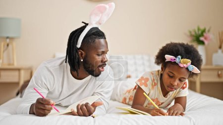 Foto de Afroamericanos padre e hija usando divertido dibujo de la diadema en el cuaderno acostado en la cama en el dormitorio - Imagen libre de derechos