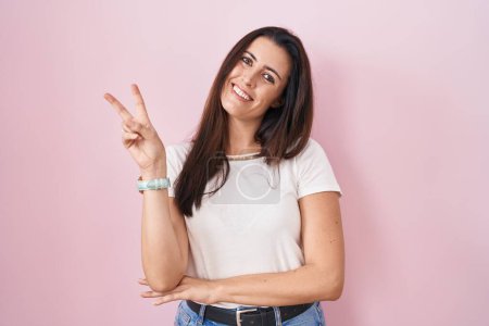 Téléchargez les photos : Jeune femme brune debout sur fond rose souriant avec un visage heureux clin d'oeil à la caméra faisant signe de victoire avec les doigts. numéro deux. - en image libre de droit