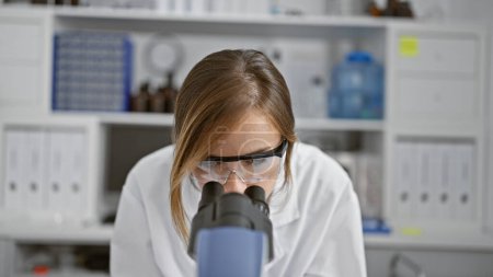 Foto de Atractiva joven científica rubia con gafas que se concentra seriamente en la investigación médica utilizando el microscopio en el laboratorio, descubriendo avances en la medicina - Imagen libre de derechos