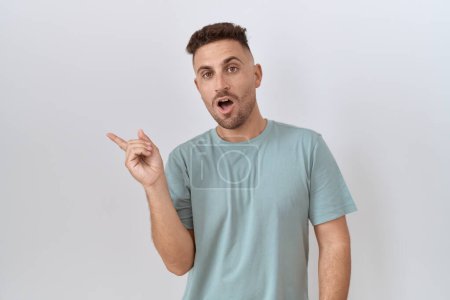 Foto de Hispanic man with beard standing over white background surprised pointing with finger to the side, open mouth amazed expression. - Imagen libre de derechos