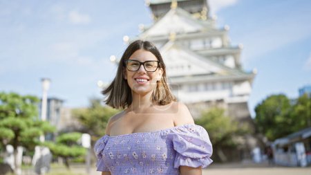 Téléchargez les photos : Beauté latine joyeuse dans les lunettes, une femme brune pose avec un sourire confiant et insouciant au célèbre château d'osaka, débordant de joie et de bonheur dans le tourisme patrimonial japonais - en image libre de droit