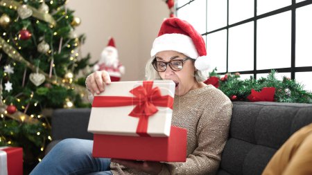 Foto de Mujer de mediana edad con cabello gris usando sombrero de Navidad desembalaje regalo con la cara sorprendida en casa - Imagen libre de derechos