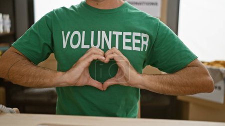 Photo for Hispanic male volunteer, hands lovingly working with heart gesture at community charity center, symbolizing support and altruism amidst donation boxes, wearing uniform t-shirt, indoor view - Royalty Free Image
