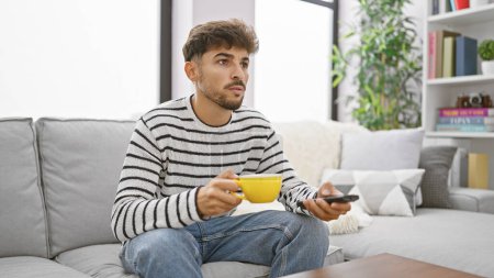 Photo for Young arab man watching television drinking coffee at home - Royalty Free Image
