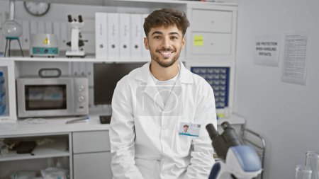 Téléchargez les photos : Un jeune homme arabe confiant, un scientifique souriant, plonge dans la recherche dans son laboratoire, perfectionnant son métier avec un microscope et une immense passion pour la science - en image libre de droit