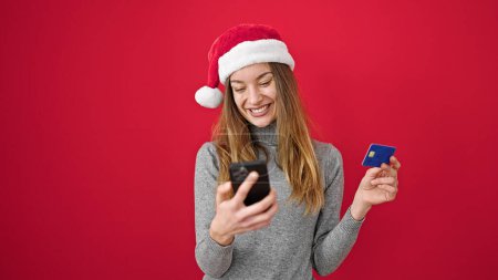 Foto de Mujer caucásica joven haciendo compras en línea de Navidad con teléfono inteligente sobre fondo rojo aislado - Imagen libre de derechos