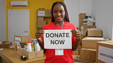 Téléchargez les photos : Belle femme afro-américaine tressée souriant avec confiance alors qu'elle se porte volontaire. soutenir un don maintenant message dans un centre de charité, montrant l'unité et l'altruisme à travers son service. - en image libre de droit