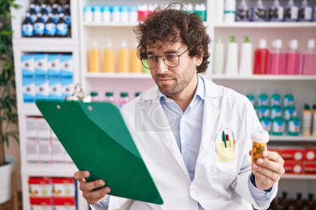 Photo for Young hispanic man pharmacist holding pills bottle reading document at pharmacy - Royalty Free Image