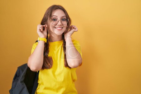 Foto de Mujer caucásica joven con mochila de estudiante sobre fondo amarillo sonriendo tirando de las orejas con los dedos, gesto divertido. problema de audición - Imagen libre de derechos
