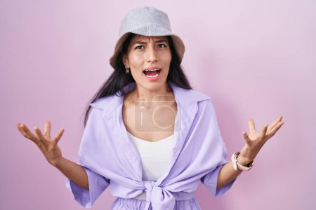 Foto de Mujer hispana joven de pie sobre fondo rosa usando sombrero loco y loco gritando y gritando con expresión agresiva y los brazos levantados. concepto de frustración. - Imagen libre de derechos