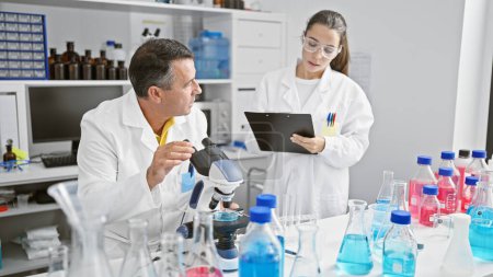 Photo for Male and female scientists working together in lab, serious concentration on their research, writing on clipboard, using microscope - Royalty Free Image