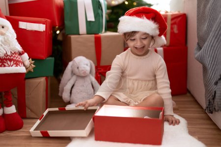Photo for Adorable girl unpacking gift sitting by christmas tree at home - Royalty Free Image