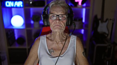 Photo for Serious grey-haired senior woman streamer engrossed in a nighttime digital game at home, relaxed yet focused, sitting with headset and gamepad in her gaming room - Royalty Free Image