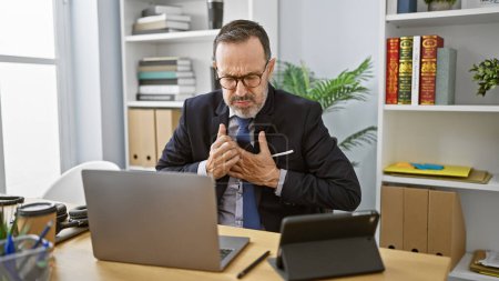 Photo for Stressed middle-aged man with grey hair, working hard in business, coughing at his office desk despite illness - Royalty Free Image
