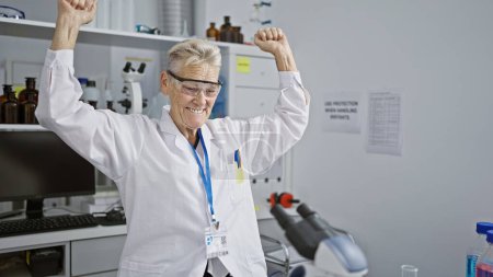 Téléchargez les photos : Célébrant une victoire, une scientifique chevelue aux cheveux gris sourit tout en travaillant au microscope de son laboratoire, débordant d'enthousiasme au cœur de la médecine de la recherche. - en image libre de droit