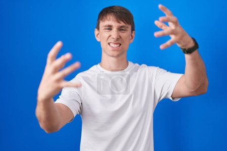 Photo for Caucasian blond man standing over blue background shouting frustrated with rage, hands trying to strangle, yelling mad - Royalty Free Image