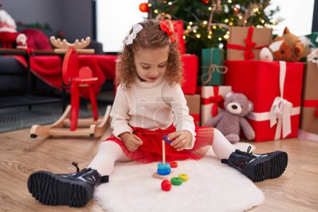 Photo for Adorable blonde girl playing with hoops toy sitting on floor by christmas tree at home - Royalty Free Image