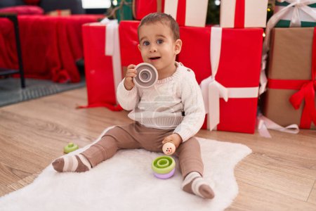 Foto de Adorable niño hispano sosteniendo maraca sentada en el suelo por regalos de Navidad en casa - Imagen libre de derechos