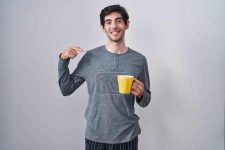 Photo for Young hispanic man wearing pajama drinking a cup of coffee looking confident with smile on face, pointing oneself with fingers proud and happy. - Royalty Free Image