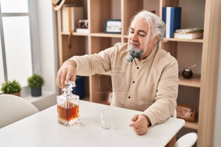 Foto de Hombre de pelo gris de mediana edad bebiendo un vaso de whisky sentado en la mesa en casa - Imagen libre de derechos