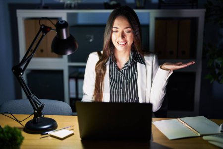 Téléchargez les photos : Jeune femme brune travaillant au bureau la nuit avec un ordinateur portable souriant présentation joyeuse et pointant la paume de la main en regardant la caméra. - en image libre de droit