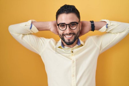 Téléchargez les photos : Jeune homme hispanique portant des vêtements et des lunettes de travail relaxant et étirant, les bras et les mains derrière la tête et le cou souriant heureux - en image libre de droit