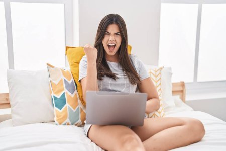 Photo for Hispanic woman using computer laptop on the bed annoyed and frustrated shouting with anger, yelling crazy with anger and hand raised - Royalty Free Image