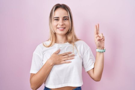Téléchargez les photos : Jeune femme blonde debout sur fond rose souriant jurant avec la main sur la poitrine et les doigts vers le haut, faisant un serment de fidélité - en image libre de droit