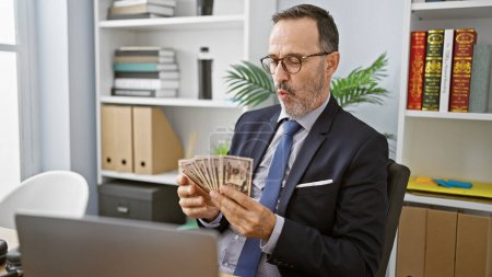 Foto de Gerente experimentado en canas y gafas, un hombre de mediana edad, sumergiéndose en un negocio de dinero serio, contando dólares en la mesa de su oficina - Imagen libre de derechos