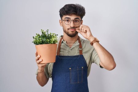 Téléchargez les photos : Homme arabe avec la barbe tenant la bouche de pot de plante verte et les lèvres fermées comme zip avec les doigts. secret et silencieux, tabou parlant - en image libre de droit