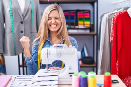 Photo for Blonde woman dressmaker designer using sew machine very happy and excited doing winner gesture with arms raised, smiling and screaming for success. celebration concept. - Royalty Free Image