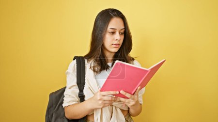 Photo for Young and beautiful hispanic female student, deeply engrossed while reading her book, standing casually against an isolated yellow backdrop with a cool backpack, portraying an attractive lifestyle - Royalty Free Image