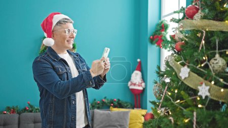 Téléchargez les photos : Jeune homme caucasien faire une photo de l'arbre de Noël par smartphone à la maison - en image libre de droit