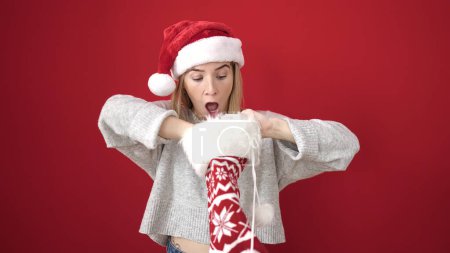 Photo for Young blonde woman wearing christmas hat looking inside of sock over isolated red background - Royalty Free Image
