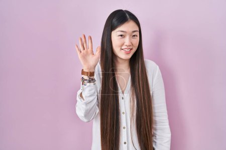 Téléchargez les photos : Jeune femme chinoise debout sur fond rose renonçant à dire bonjour heureux et souriant, geste de bienvenue amical - en image libre de droit